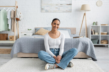 cheerful african american student in jeans sitting with crossed legs near bed at home.