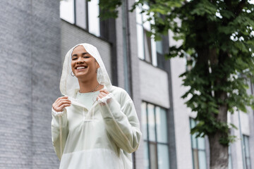 carefree african american woman in waterproof raincoat standing under rain outdoors.