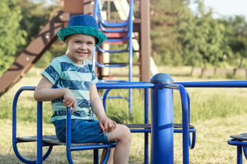 A cute boy is sitting on a merry-go-round in the playground, he is smiling and posing for the camera.