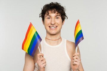 overjoyed nonbinary person in colorful beads holding small lgbt flags isolated on grey.