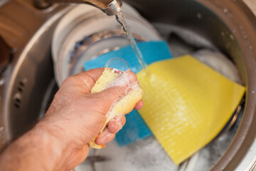 Fototapeta premium Washing dishes. A man's hand squeezes a soapy solution from a sponge, with a blue rag for washing dishes in the kitchen. An image about housework, apartment care and cleanliness.