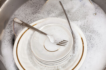 Washing dishes. Forks, spoons, plates of different sizes in the sink under running water. An image about housework, apartment care and cleanliness.
