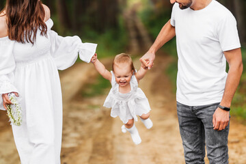 Fototapeta premium Baby swings holding hands of parents. Summer family walk.