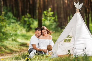 Young parents play with their little daughter in nature. People are sitting in grass near playhouse.