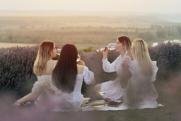 Fototapeta na wymiar Friends having picnic in the lavender field. Group of young women sitting on lavender field at sunset on summer day. Girlfriends drinking wine on outdoor party.