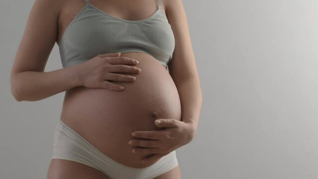 Pregnant woman wearing underwear stroking her belly. Unrecognizable expectant mother gently caressing her abdomen with baby on standing white background