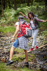 Young couple hiking in forest. Crossing a creek, boyfriend reaches out to help. Summertime, mountain landscape. Freedom, friendship, love, nature concept.