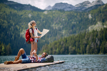 A young couple is taking a rest on the dock at the lake during mountain hiking. Trip, nature, hiking