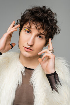 portrait of stylish pangender person in silver accessories and white faux fur jacket posing with hands near face isolated on grey.