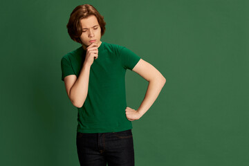 Portrait of young guy wearing casual clothes standing and thinking with unconfident facial expression over green background. Unware, doubtful, uncertain