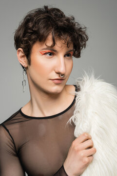 portrait of young pansexual person with makeup holding white faux fur jacket and looking away isolated on grey.