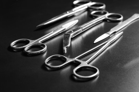 Stainless steel surgical instruments (forceps, scalpel and set of scissors) lying on a black matte surface, black & white image, selective focus, macro shot