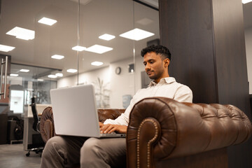 A young multiethnic businessman is sitting in a rich leather armchair and using his laptop in the office.