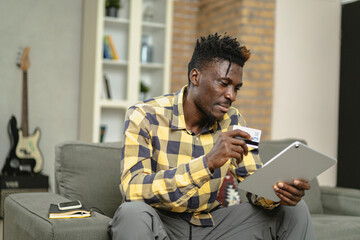 Young African man using digital tablet at home. Smiling man reading news while sitting on the couch at home.