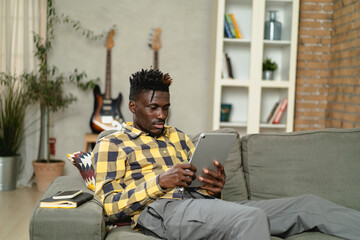 Young African man using digital tablet at home. Smiling man reading news while sitting on the couch at home.