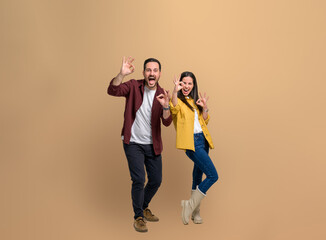 Full length of stylish young couple showing OK Okay signs and screaming ecstatically while looking at camera. Excited man and woman gesturing while posing happily over beige background