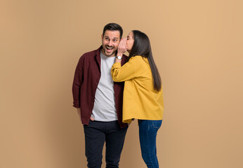 Young girlfriend whispering secret into shocked boyfriend's ear. Couple dressed in casual gossiping while standing together isolated against beige background - obrazy, fototapety, plakaty