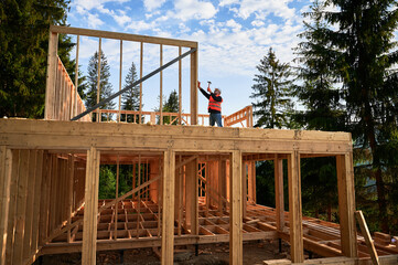 Wooden two-story, frame-built home is being erected by the carpenter near the forest. Man is driving in nails with hammer. Underpinning idea of this venture is modern, ecological building practices.