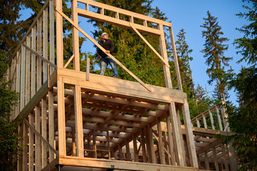 Fototapeta na wymiar Carpenter constructing wooden framed two-story house near the forest. Bearded man with glasses holds hammer, dressed in protective helmet. Concept of environmentally friendly modern construction.
