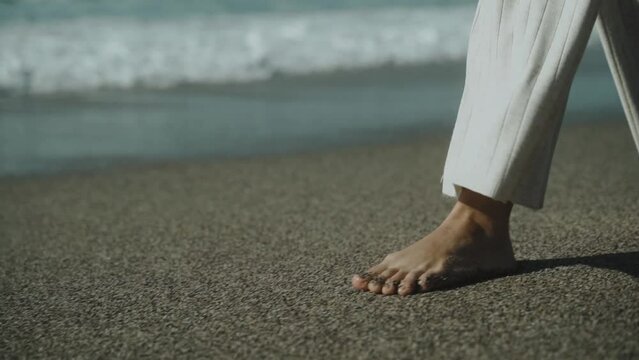 Slender woman's legs walking along the sandy beach. White developing pants.