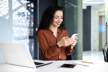 Beautiful and successful business woman with curly hair at workplace inside office using phone, latin american boss typing messages and browsing internet pages online smiling contentedly.