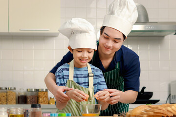 Happy smiling Asian father and son kid with apron and chef hat cooking meal together at kitchen,...