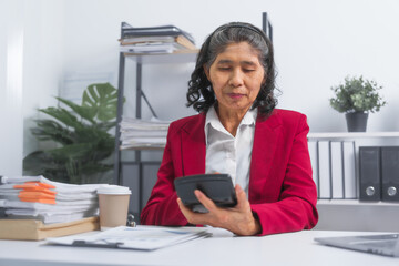 Confident asian elderly businesswoman in red suit working at desk planning bookkeeping project, thinking and reading, sitting at workplace.