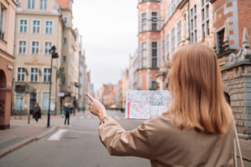 Back view of attractive young female tourist is exploring new city. Woman holding a paper map on central square in Gdansk old town in Poland. High quality photo