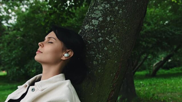 Woman listening to music on wireless headphones with her eyes closed in park. Female musician uses earbuds leaning against tree trunk outdoors