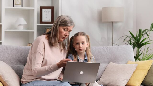 Grayhaired grandmother and little granddaughter are sitting on the sofa in the living room, resting and watching funny video cartoons on the laptop, talking, having fun.