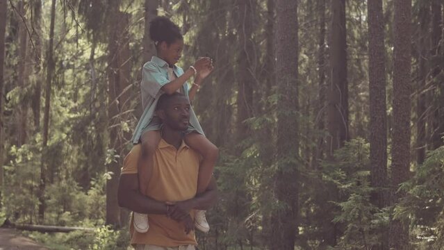 Medium slowmo of African American little girl taking photos on smartphone sitting on dad shoulders while spending day together in forest at summertime