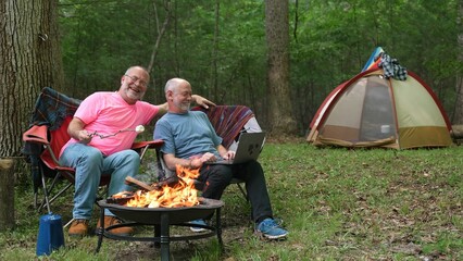 Two gay men with pride flag and tent in tent talking to laptop on video chat.