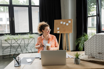 happy architectural designer in glasses holding coffee to go near laptop and residential house model in office.