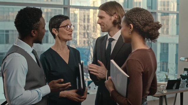 Medium shot of four professional multiethnic lawyers standing in contemporary office with big windows having conversation