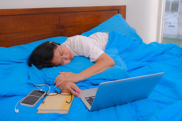 A woman in a white t-shirt sleeping on her stomach; a laptop, a book and a cell phone next to her.