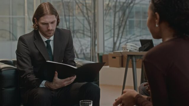 Medium shot of confident young Caucasian businessman in elegant suit and tie reading contract during negotiations with partners in contemporary office