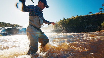 Fly fishing in a rapid river. Fisherman in waders slowly walks from the furious murky river