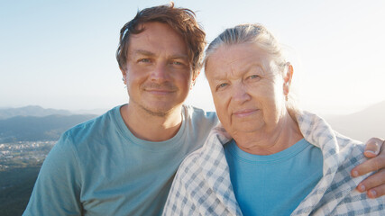 Portrait of young man and elderly woman. Young man hugs senior caucasian woman outdoor at sunrise. Models look into the camera