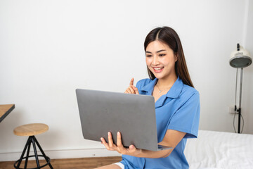 Young Asian woman with beautiful face, long hair in blue shirt sitting on white bedroom bed at home with cup of coffee and video call with laptop talking to relatives on holiday. vacation concept.