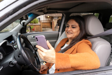 Fototapeta na wymiar Confident smiling mature Indian woman holding mobile phone, checking email sitting inside new car. Transportation, distracted driving concept