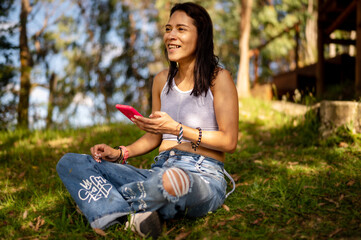 Happy woman sitting on grass in park holding a smart phone, happy smiling woman outside