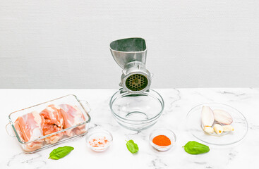 Manual meat grinder with glass bowl,boneless chicken thighs,peeled onions,garlic on a marble table .