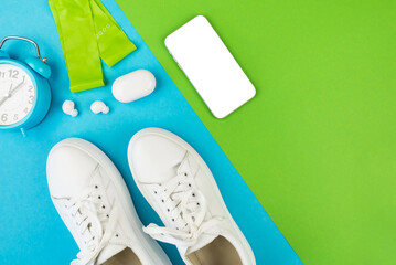 Top view photo of white sneakers, smart phone with white screen, headphones and alarm clock on blue green background with copy space.