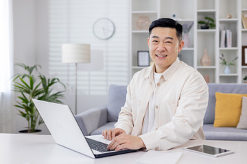Fototapeta na wymiar Portrait of successful Asian programmer, man working remotely from home office, businessman smiling and looking at camera using laptop at work sitting in living room at home.