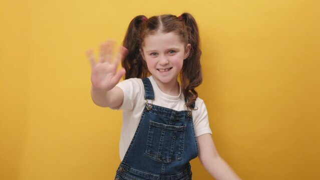 Portrait of happy little girl kid smiling friendly at camera and waving hands gesturing hello or goodbye, welcoming with hospitable expression, posing isolated on yellow color background in studio