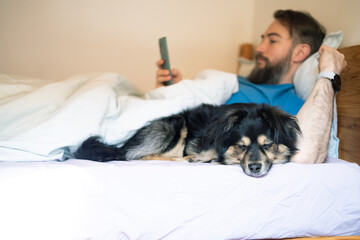Man in a blue shirt lying on a bed with a dog and using a smartphone.