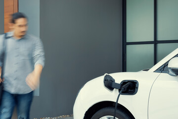 Focus electric car charging at home charging station with blurred progressive man walking in the...