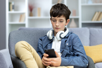 Serious child of a teenage boy in headphones sitting at home on the sofa, using the phone. Listens to music, watches videos. He chats, sits on social networks, plays online games. © Liubomir