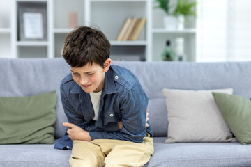 A child, a boy, a teenager, sits alone at home on the sofa and holds his stomach. Suffering from...