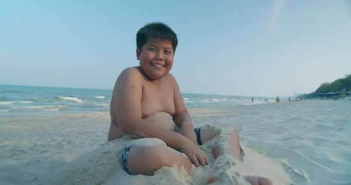 (Slow motion) A happy Asian boy who had a rather chubby figure, enjoying and playing in the sand on the sea beach in the evening sunset time.
